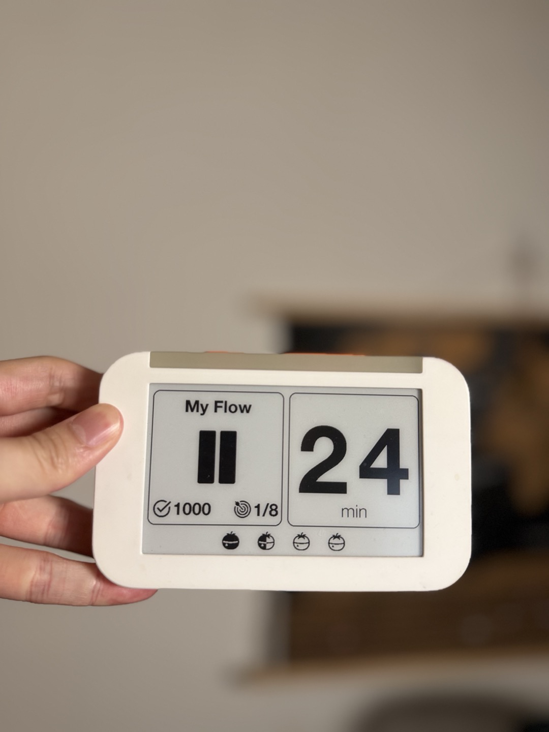 手持白色心流鐘 Flow Clock 實體番茄鐘，電子紙螢幕顯示生涯累積 1000 顆番茄鐘、今日進度 1/8、剩餘 24 分鐘